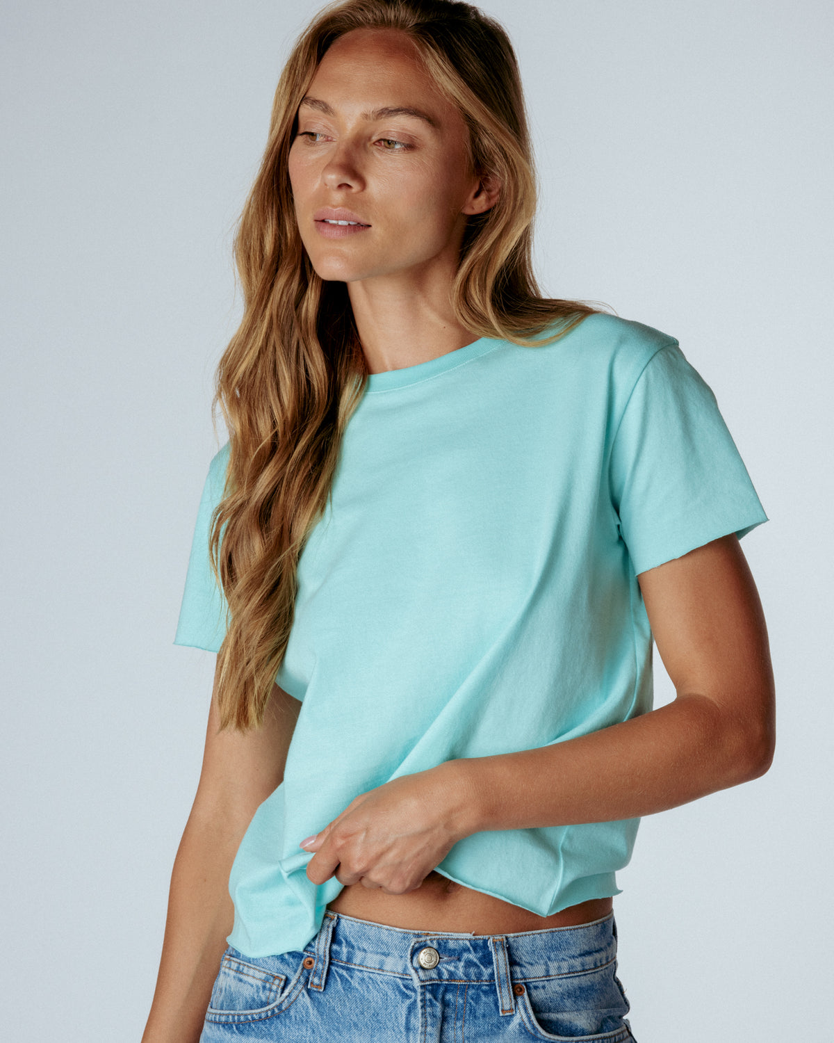 A woman wearing a light mint green short-sleeve t-shirt and blue jeans, photographed against a neutral light gray studio background.