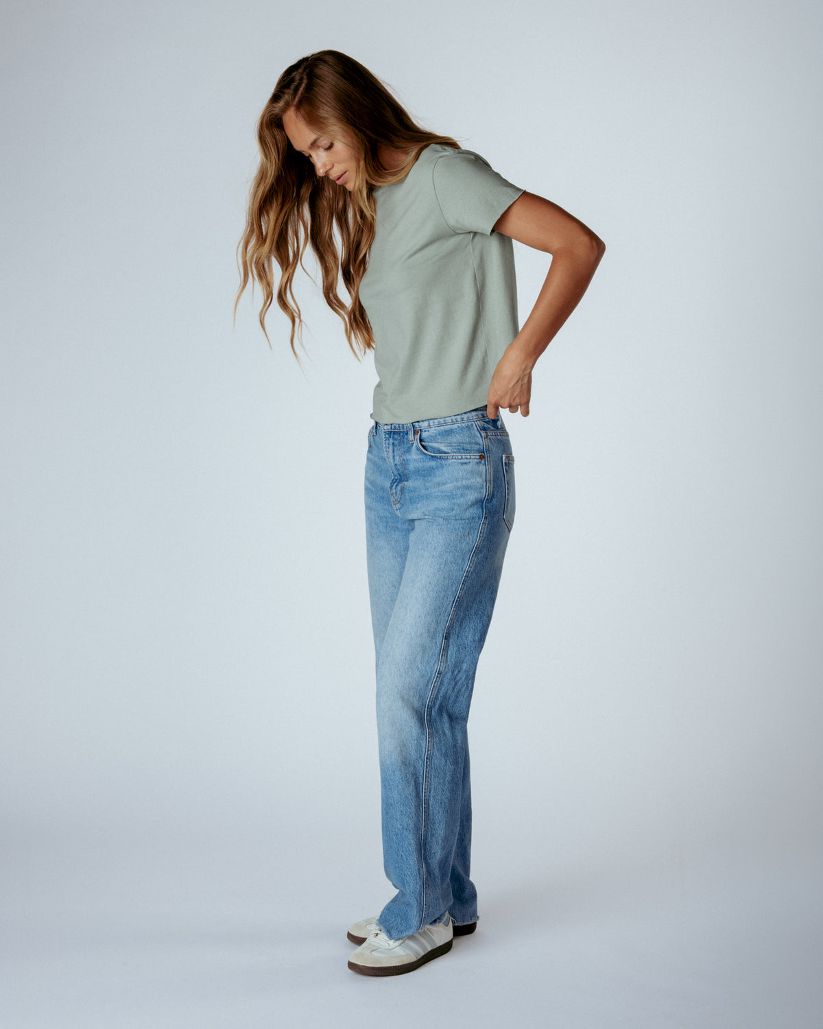 A woman modeling a light sage green short-sleeve t-shirt and light-wash blue jeans, standing against a light gray studio background.