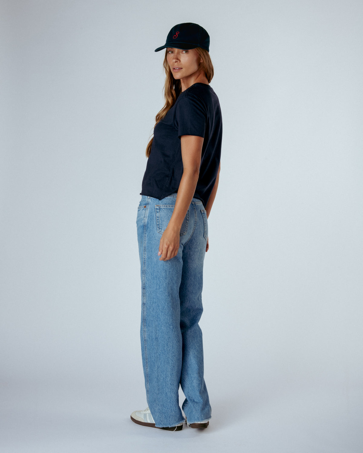A woman stands in profile wearing a black short-sleeve t-shirt, light blue wide-leg jeans, white shoes, and a black baseball cap against a light gray studio background.