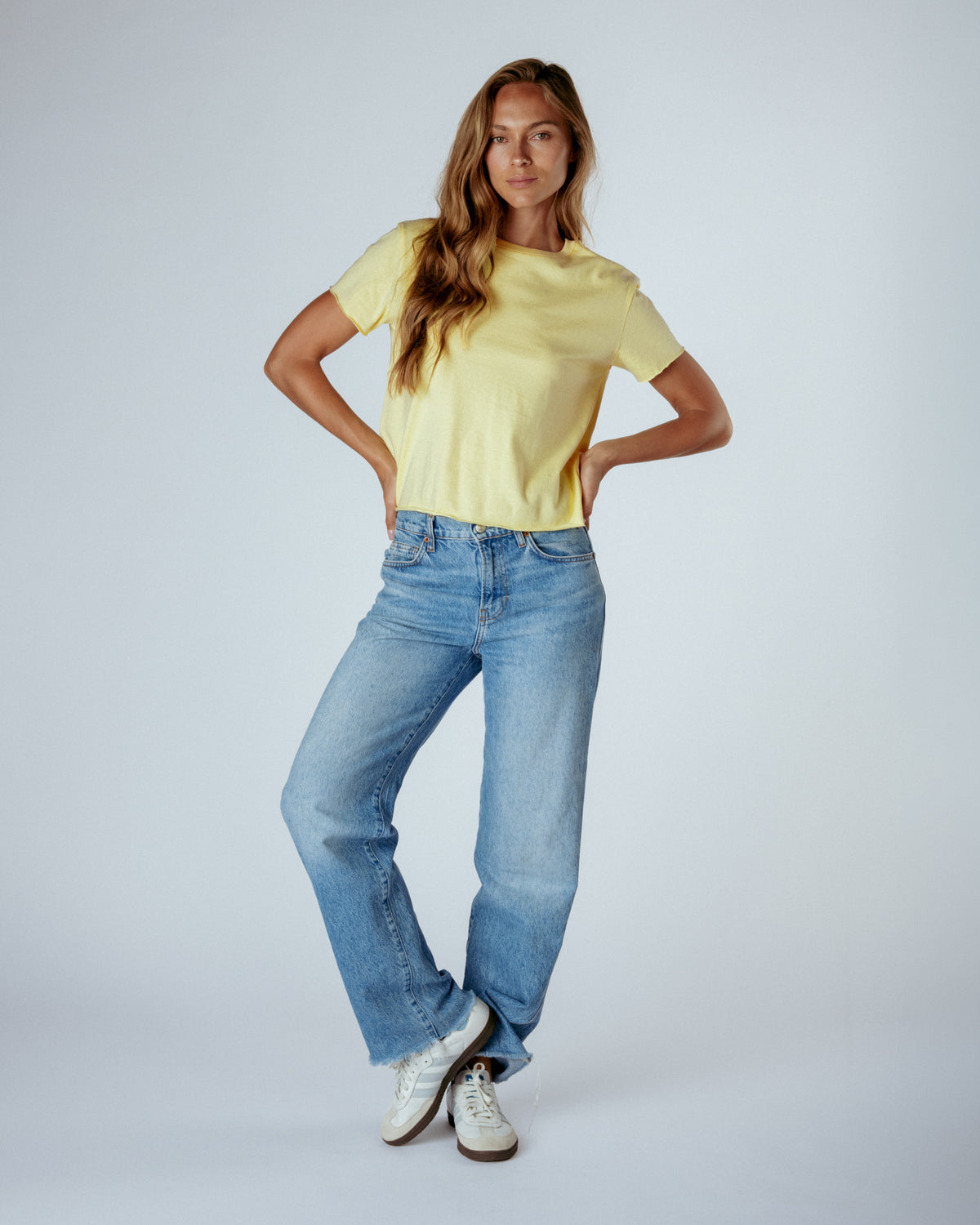 A woman modeling a pale yellow short-sleeve crewneck t-shirt and light-wash blue straight-leg jeans, paired with white sneakers, against a light gray studio background.