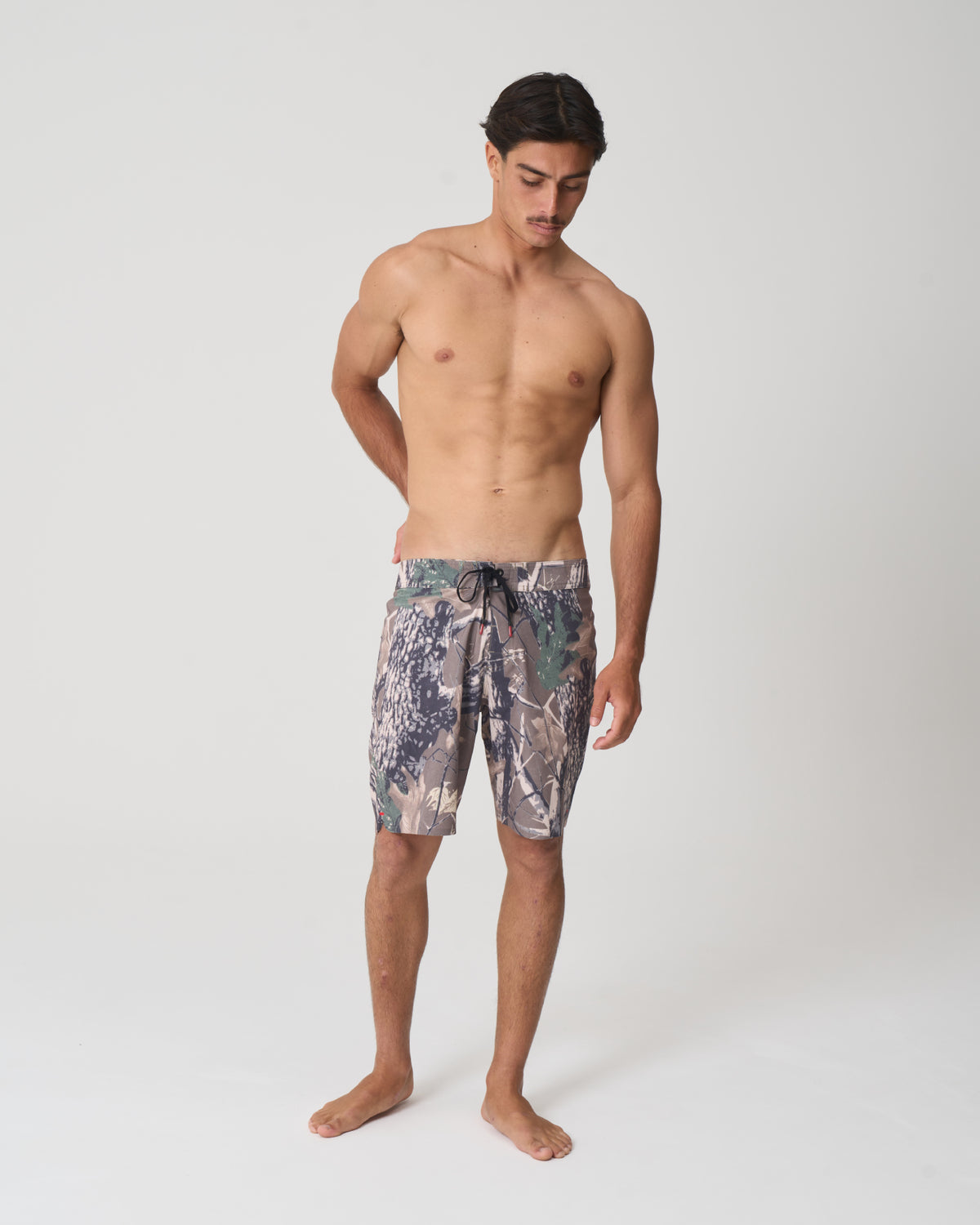 A shirtless male model stands barefoot against a light gray studio background, wearing patterned swim trunks with a camouflage-like design in muted green, gray, beige, and blue tones.