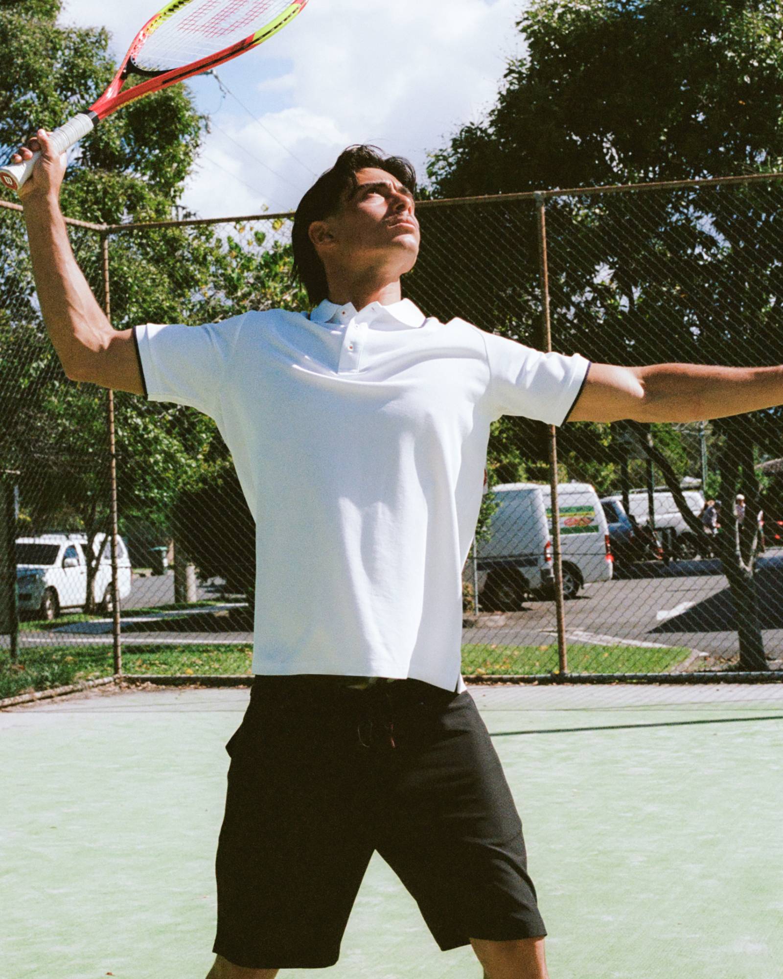Man playing tennis on a court with trees and vehicles in the background