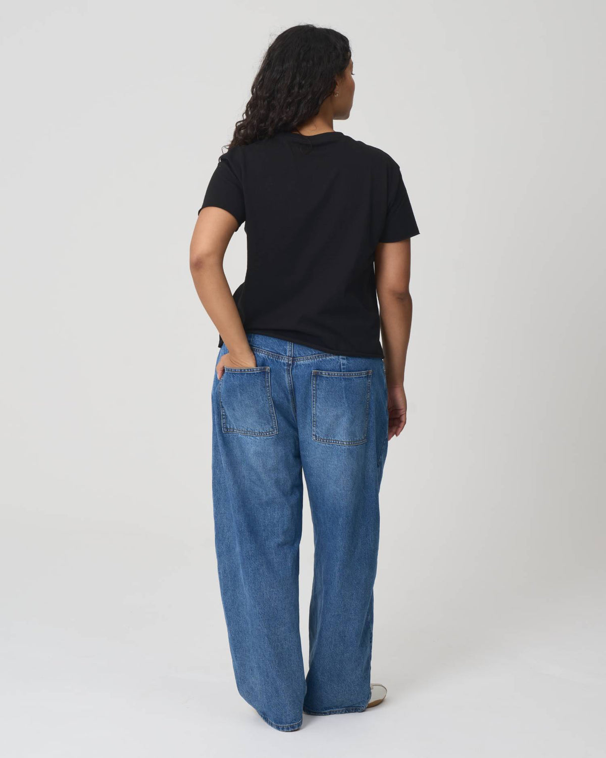 A model shown from the back wearing a black short-sleeve t-shirt and wide-leg blue denim jeans, standing against a light gray studio background.